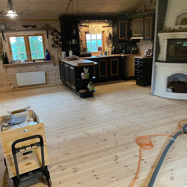 Koselig kjøkken i en hytte, med panelvegger, lyst tregulv og en hvit peisovn til høyre, samt mørke skap og arbeidsbenk.